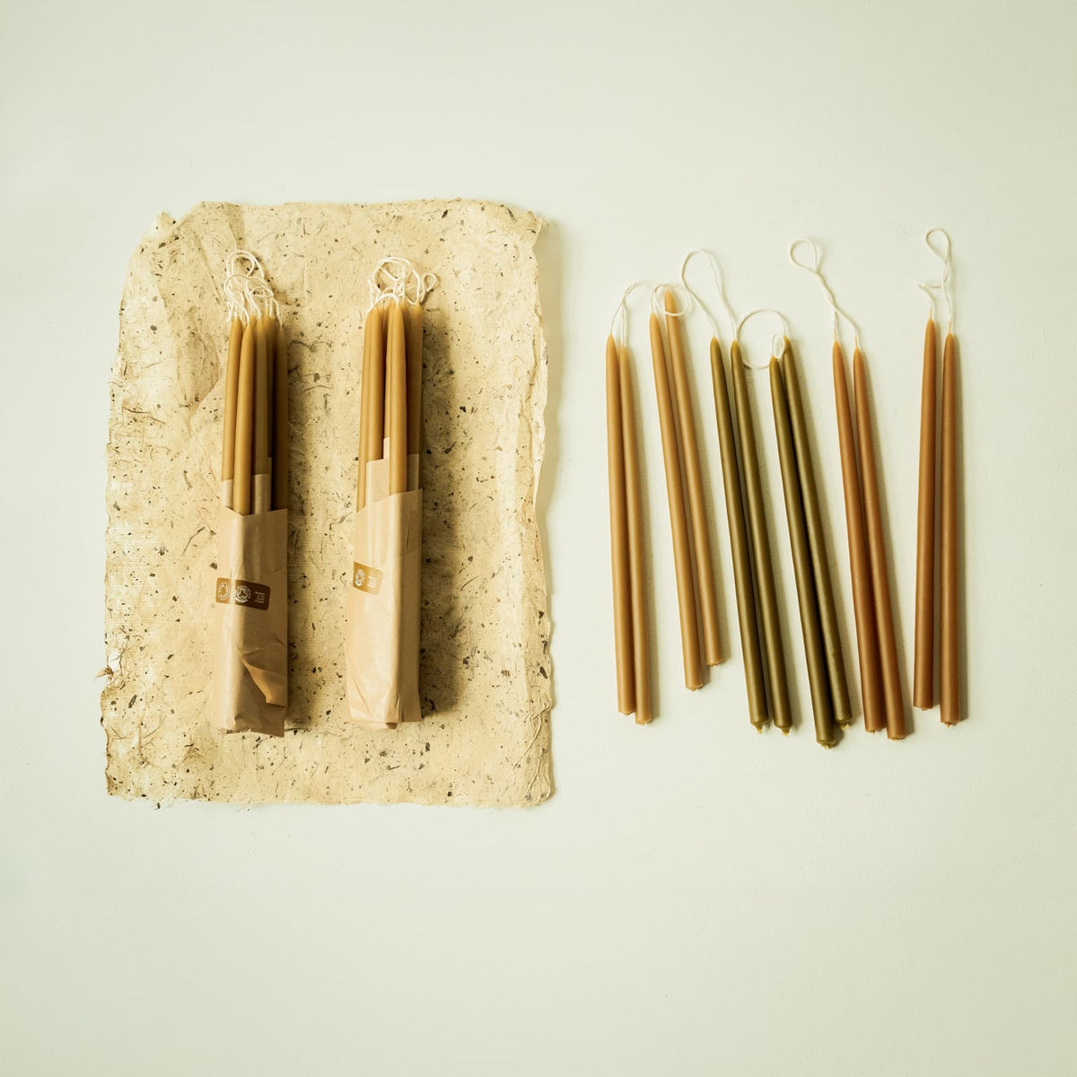 Two wrapped packs of natural beeswax slim dinner candles beside twelve loose companion candles in mixed colours, on handmade paper